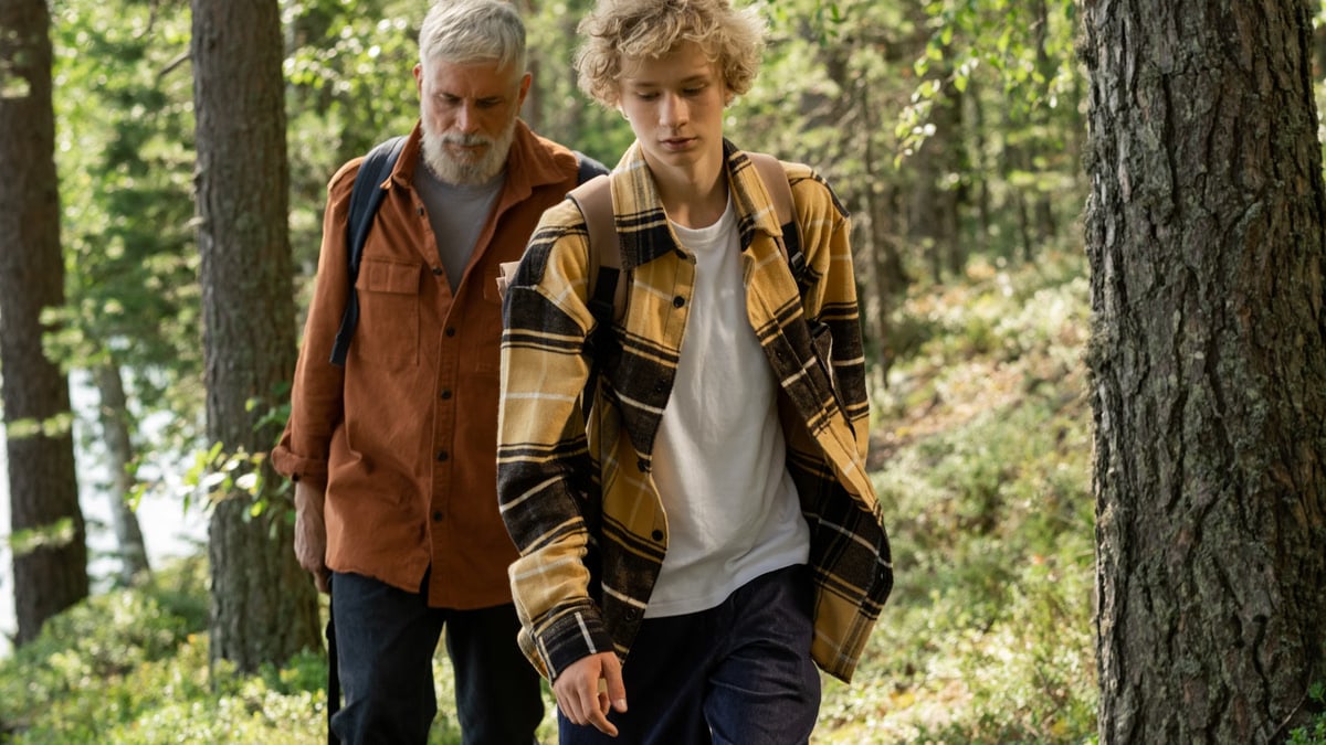 An elderly man and a teenage boy hiking through a lush green forest, enjoying nature.